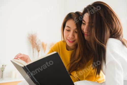 Preview: lesbian happy couple sitting on bed reading book together near window in bedroom.