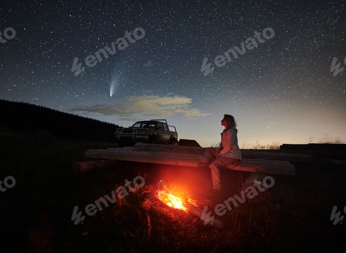 Preview: Female traveler sitting near campfire under night starry sky.