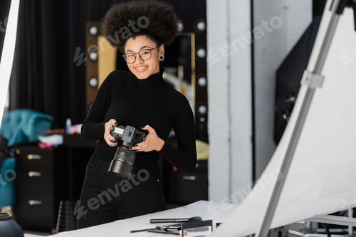 Preview: cheerful african american content producer with digital camera smiling near decorative cosmetics on