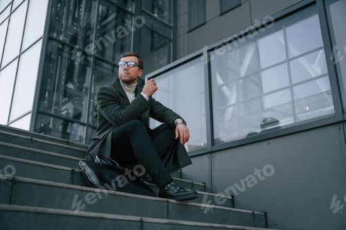 Vista previa: fumando. Un hombre guapo con ropa formal está al aire libre cerca del edificio de negocios