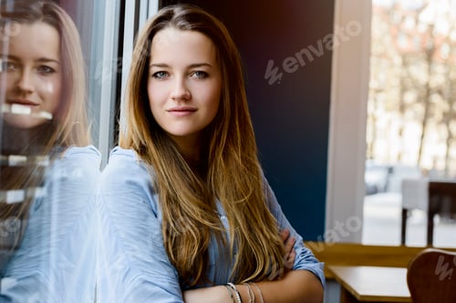 Preview: Young woman leaning against window arms folded looking at camera smiling