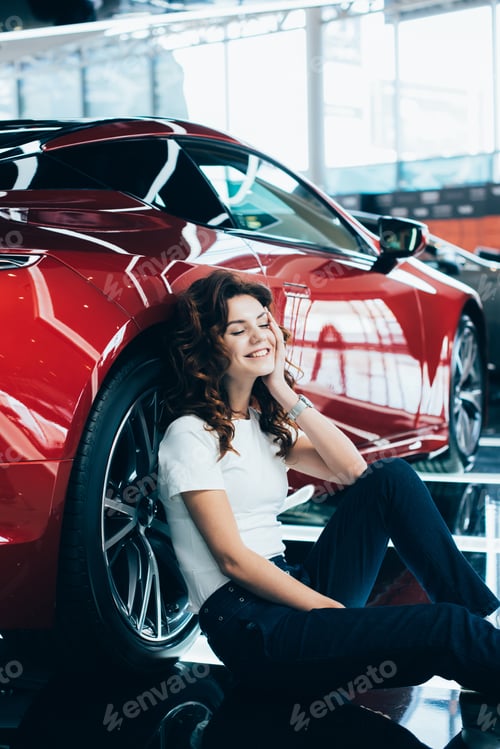 Preview: selective focus of happy woman with closed eyes sitting on floor near new red car