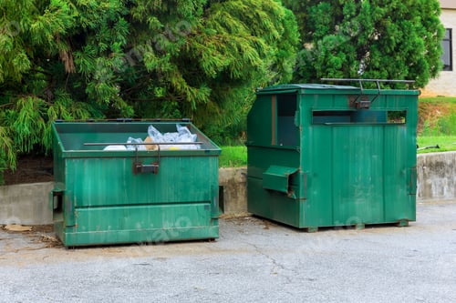 Preview: Two green dumpsters positioned beside concrete wall in parking area surrounded by trees on clear day