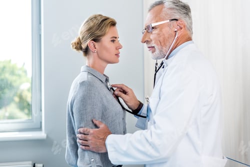 Preview: side view of mature male doctor examining female patient by stethoscope in hospital room