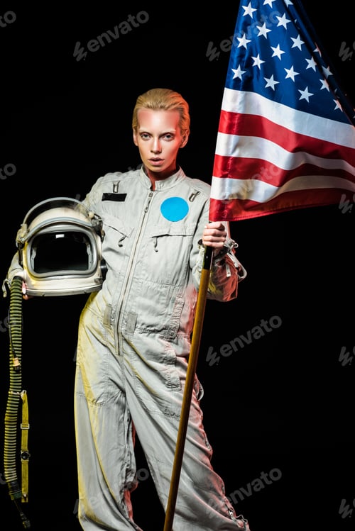 Preview: beautiful female cosmonaut in spacesuit holding helmet and usa flag, isolated on black