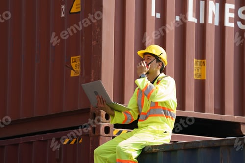 Preview: Worker checks laptop while sitting on container at shipping yard during daylight hours
