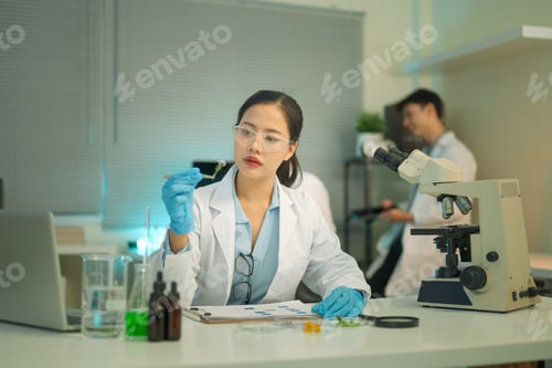 Preview: Female scientist records results while examining plant samples in petri dishes beside a microscope