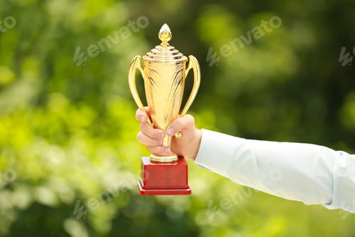 Preview: Young businessman holding gold trophy cup in green park, closeup