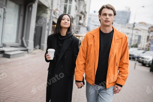 Preview: Stylish man holding hand of asian girlfriend with coffee to go on urban street