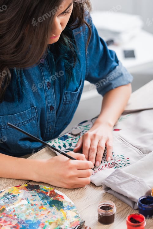 Preview: focused female fashion designer painting on jacket at working table in clothing design studio