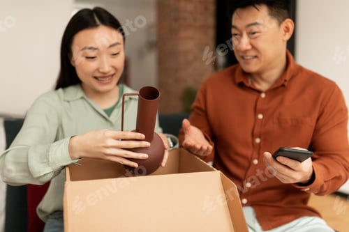Preview: Contented asian spouses unpacking box after successful online shopping, sitting on sofa and smiling