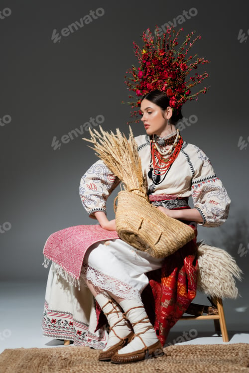 Preview: full length of young ukrainian woman in red wreath with flowers holding bag with wheat on dark grey