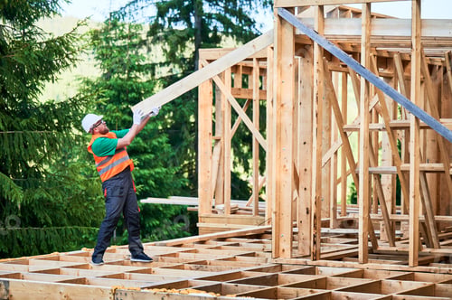 Vista previa: Carpenter holding large beam while builds wooden frame house near the forest.