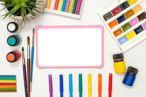 Preview: Childrens pink digital tablet with a blank screen on a white table with multicolor drawing supplies.