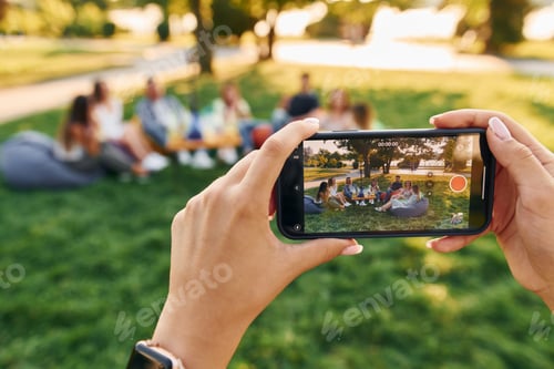 Preview: Making photo by using smartphone. Group of young people have a party in the park at summer daytime