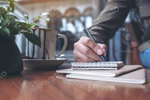 Preview: Closeup image of a woman's hand writing on blank notebook with coffee cup on wooden table