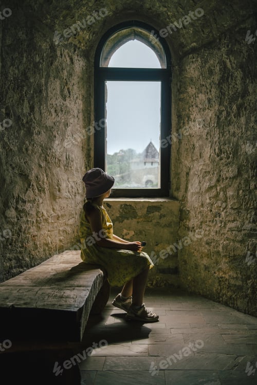 Preview: Teen girl wearing dress and sun hat sitting on wooden bench in medieval castle and looking to window