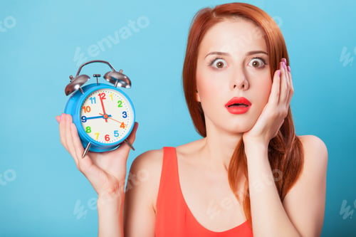 Preview: Surprised redhead women woth alarm clock on blue background.