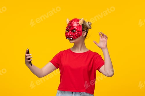 Preview: young female wearing scary devil mask using her phone yellow background halloween