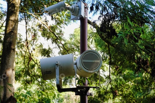 Preview: Pole with a camera and loudspeakers surrounded with green trees in the park