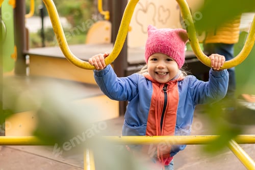 Preview: A cheerful smiling little girl in blue jacket and pink hat playing on modern playground in city park