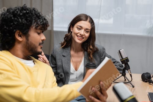 Preview: curly indian radio host in yellow jumper showing notebook to happy brunette colleague in grey
