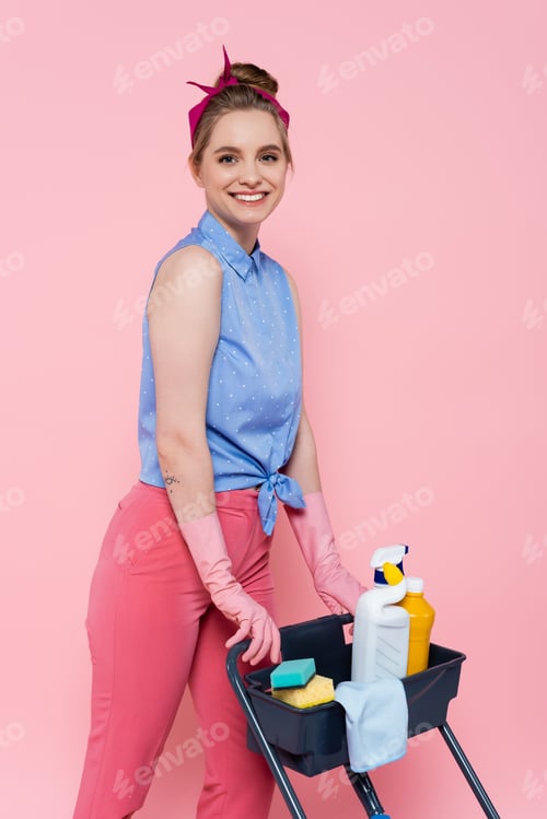 Preview: happy young woman in rubber gloves standing near housekeeping cart with cleaning supplies isolated