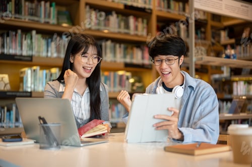Preview: Glasses asian man student and woman friend looking at tablet and making a fist at table in library.