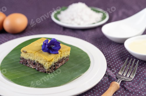 Preview: Black sticky rice and custard on a banana leaf in a white plate with butterfly pea flowers.