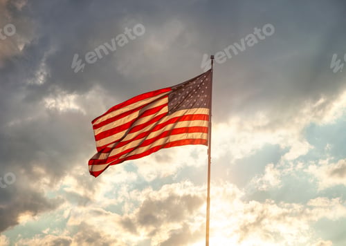 Preview: Back lit American flag waving under clouds with sun rays at sunset
