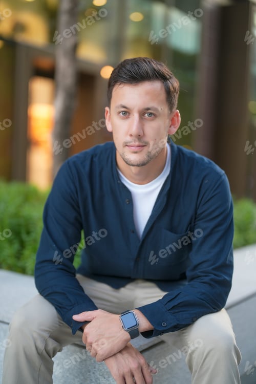 Preview: Young man sits casually outdoors in urban setting during the day