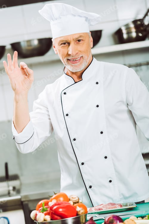 Preview: handsome smiling male chef in uniform looking at camera and showing ok sign in restaurant kitchen