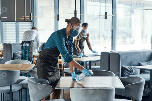 Preview: Busy young male waiters in protective workwear cleaning tables in restaurant