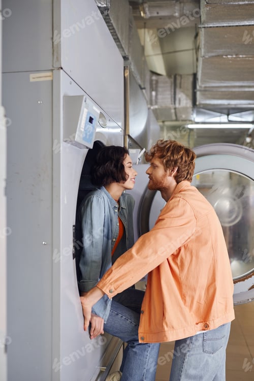 Preview: side view of smiling asian woman sitting on washing machine near romantic boyfriend in coin laundry