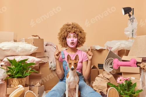 Preview: Stunned curly haired woman points above shows something shocking applies beauty mask on face dressed