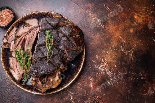Preview: Baked and sliced Venison leg. roast meat of deer with bone on a plate. brown background. top view