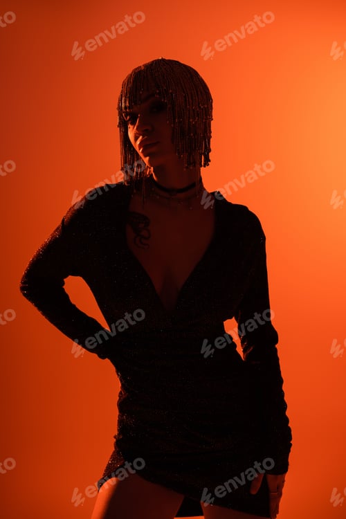 Preview: tattooed woman in black dress and metallic headwear posing with hand on waist orange background