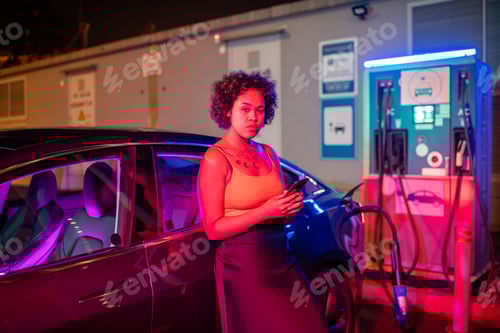 Preview: Pretty girl with smartphone standing by electric car