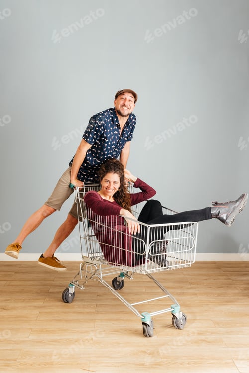 Preview: Man Pushing Woman in a Shopping Cart Indoors, Displaying Joy and Playfulness
