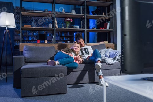 Preview: young multiracial couple with popcorn watching film on sofa together at home
