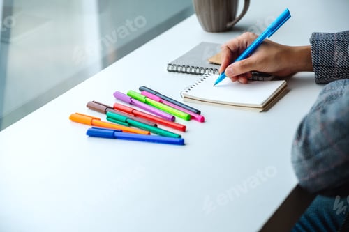 Preview: Closeup image of a woman writing on a blank notebook with colored pens and coffee cup on the table