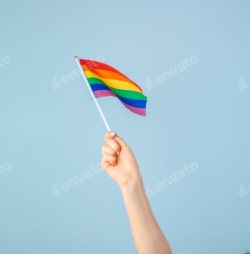 Preview: Closeup of a hand waving a small rainbow flag against a light blue background
