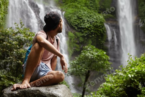 Preview: Nature, wildlife and travel concept. Young barefooted hiker wearing snapback sitting on big stone an