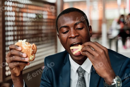 Preview: Happy young african american man having hamburger and french fries in fast food restaurant