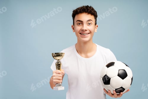 Preview: Portrait excited teenager football player with dental braces holding trophy cup and soccer ball