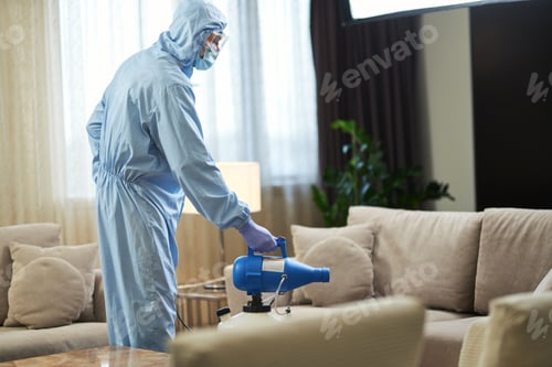 Preview: Man standing near the sofa and using disinfectant in hotel room