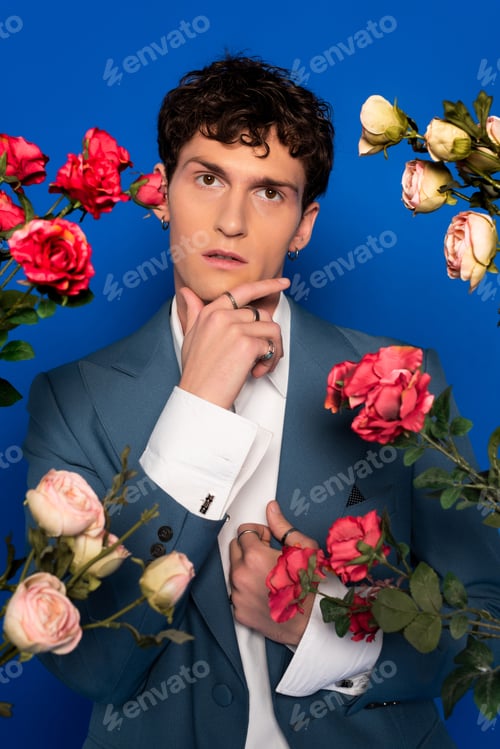 Preview: Curly man in stylish outfit touching chin near blooming flowers on blue background