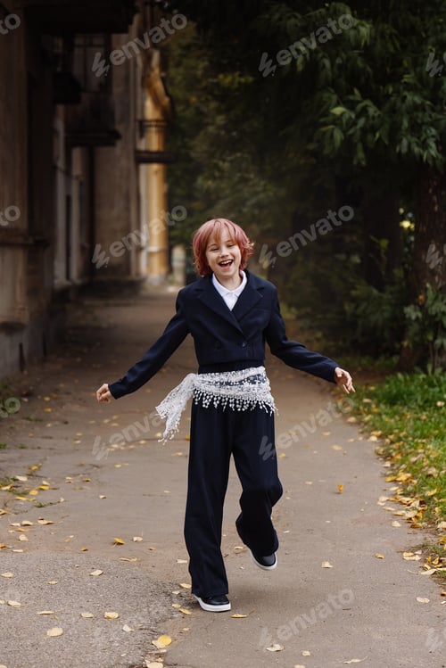 Preview: Happy schoolgirl with pink hair in navy uniform running outdoors near old building, front view