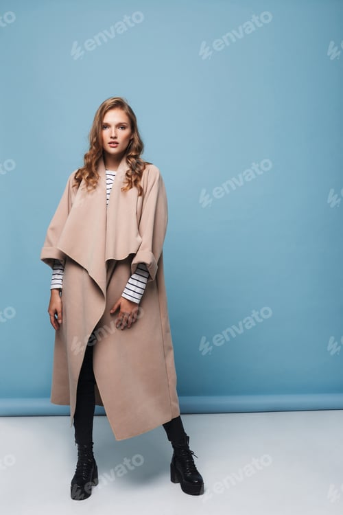 Preview: Pretty thoughtful girl with wavy hair in beige coat dreamily looking in camera over blue background