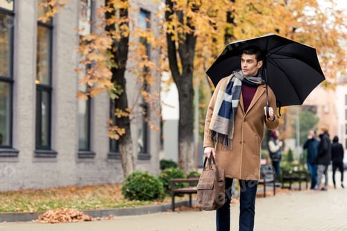 Preview: handsome stylish man in coat and scarf with umbrella walking by autumnal street